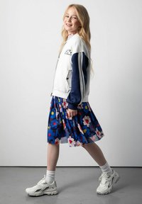 Biała kurtka bombowiec z granatowymi rękawami, granatowa spódnica w kwiatowy wzór, białe sneakersy i skarpetki do kostek, pokazana w pozycji stojącej na neutralnym tle.