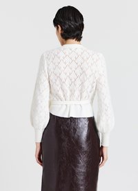 Cardigan bianco lavorato a maglia con un motivo a diamante, maniche a sbuffo e vita cintrata. Indossato sopra una gonna bordeaux lucida.