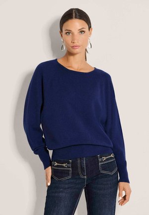 Frau mit dunklem Haar in einem tiefen Pferdeschwanz trägt einen dunkelblauen Pullover und dunkle Jeans mit goldfarbenen Kettendetails an den Taschen und posiert vor einer hellen Wand.