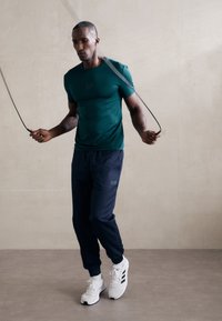 Sport t-shirt in teal with a fitted design and subtle branding, paired with navy joggers and white athletic shoes, holding a jump rope.