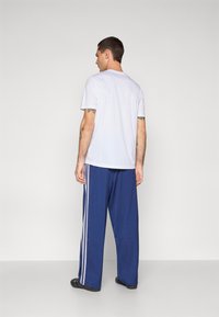 Bijela majica kratkih rukava i plave jogger hlače s bijelim prugama sa strane. Tkanina izgleda glatko; hlače su široke i opuštenog kroja.