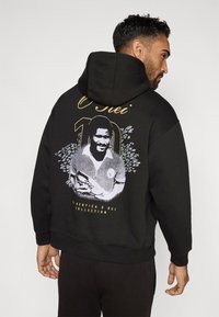 Crna dukserica s velikim grafičkim printom na leđima koja prikazuje figuru, tekst "O Rei" i "10," te cvjetni uzorak s sivim i zlatnim naglascima.