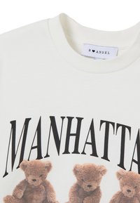 T-shirt bianco in cotone con la parola "MANHATTAN" in testo nero. Il design frontale include tre illustrazioni di orsetti in toni di marrone.