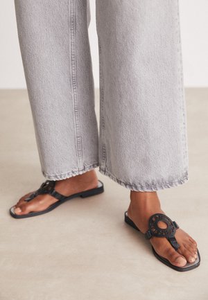 Personne portant un jean gris clair à jambes larges et des sandales noires à bout ouvert avec un motif de découpe circulaire, debout sur un sol neutre.