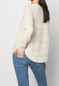 Maglione in maglia beige con texture a righe orizzontali, scollo tondo e spacchi laterali, abbinato a jeans in denim blu.