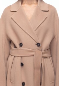 Cappotto in misto lana beige con design doppiopetto, rever a punto, bottoni neri e cintura annodata. Tessuto liscio con tasche laterali.