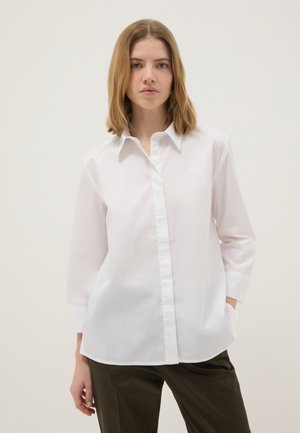 Jeune femme aux cheveux blonds mi-longs portant une chemise blanche à manches longues boutonnée et un pantalon foncé sur un fond uni.
