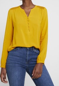 Camicia gialla a maniche lunghe con scollatura a V, dettagli in bottone e spalle con texture. Abbinata a jeans blu aderenti. Tessuto morbido.