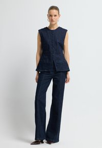 Džínová bezrukávová blůza s kulatým výstřihem, předním zapínáním na knoflíky a dvěma bočními kapsami. V kombinaci s odpovídajícími džínovými kalhotami rovného střihu.