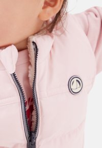 Ljusrosa vadderad väst med dragkedja, mjukt fleecefoder och en rund logotypmärke. Har huva och kontrasterande mörk dragkedja.