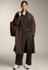 Hnedá zipsová bunda v oversize strihu, zodpovedajúce nohavice a štruktúrovaná bordó taška; biele tenisky dotvárajú outfit.