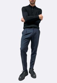 Polo nera a maniche lunghe con abbottonatura a tre bottoni, abbinata a pantaloni sartoriali blu scuro e sneakers nere. Tessuto liscio, vestibilità casual.