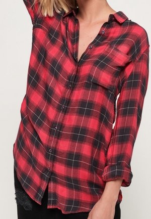 Personne portant une chemise à carreaux rouges et noirs avec des manches retroussées et un pantalon noir sur un fond uni.