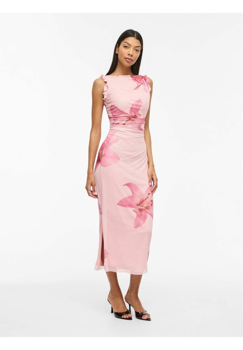 Femme portant une robe midi rose pâle sans manches avec de grands imprimés floraux, une fente latérale et des talons ouverts noirs, debout sur un fond blanc.