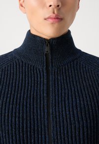 Jersey de punto azul marino con cremallera y cuello alto; tejido texturizado con tonos oscuros y claros de azul en contraste; detalle de cremallera en la parte delantera.