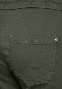 Olijfgroene broek met een gladde textuur, voorzien van een achterzak met een zilveren knop en schuine naaddetails.