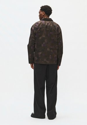 Mann steht mit dem Rücken zur Kamera, trägt eine dunkel gemusterte Jacke und weite schwarze Nadelstreifenhosen vor schlichtem weißem Hintergrund.