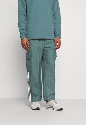 Man wearing teal cargo pants with white side stripes, matching teal long-sleeve top, and white sneakers standing against a plain background.