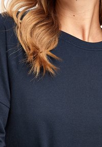 Haut à manches longues bleu marine avec un col rond et un tissu doux et texturé, présentant des coutures apparentes et une coupe décontractée. Des cheveux tombent doucement sur le vêtement.