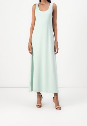 Femme portant une robe longue sans manches vert pâle avec un décolleté arrondi et des sandales à talons hauts beiges à bride cheville sur fond blanc.