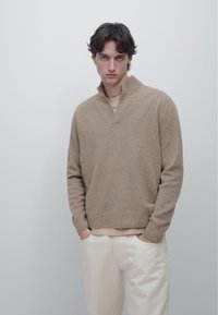 Beige strikket pullover med en h øj hals og ribbede manchetter, båret over en lys beige skjorte, kombineret med off-white bukser. Tekstureret stof.
