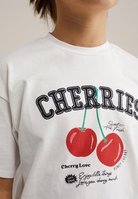 Balta medvilnė marškinėliai trumpomis rankovėmis, su raudonų vyšnių grafika, juodu siuvinėtų tekstu ir spalvingais spaudos akcentais.