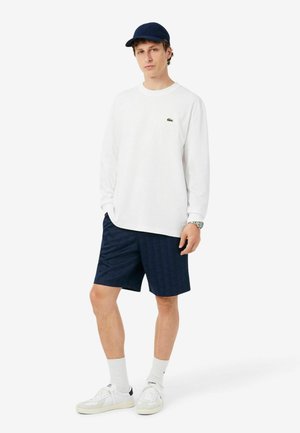 Giovane uomo che indossa una camicia bianca a maniche lunghe, pantaloncini blu navy, calzini bianchi, scarpe da ginnastica bianche, un cappellino blu navy e un orologio da polso, in piedi su sfondo bianco.