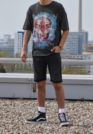 Camiseta gráfica negra con una gran cara de tigre y un fondo de remolinos coloridos; combinada con unos pantalones cortos de mezclilla negra desgastados y unas zapatillas altas negras.