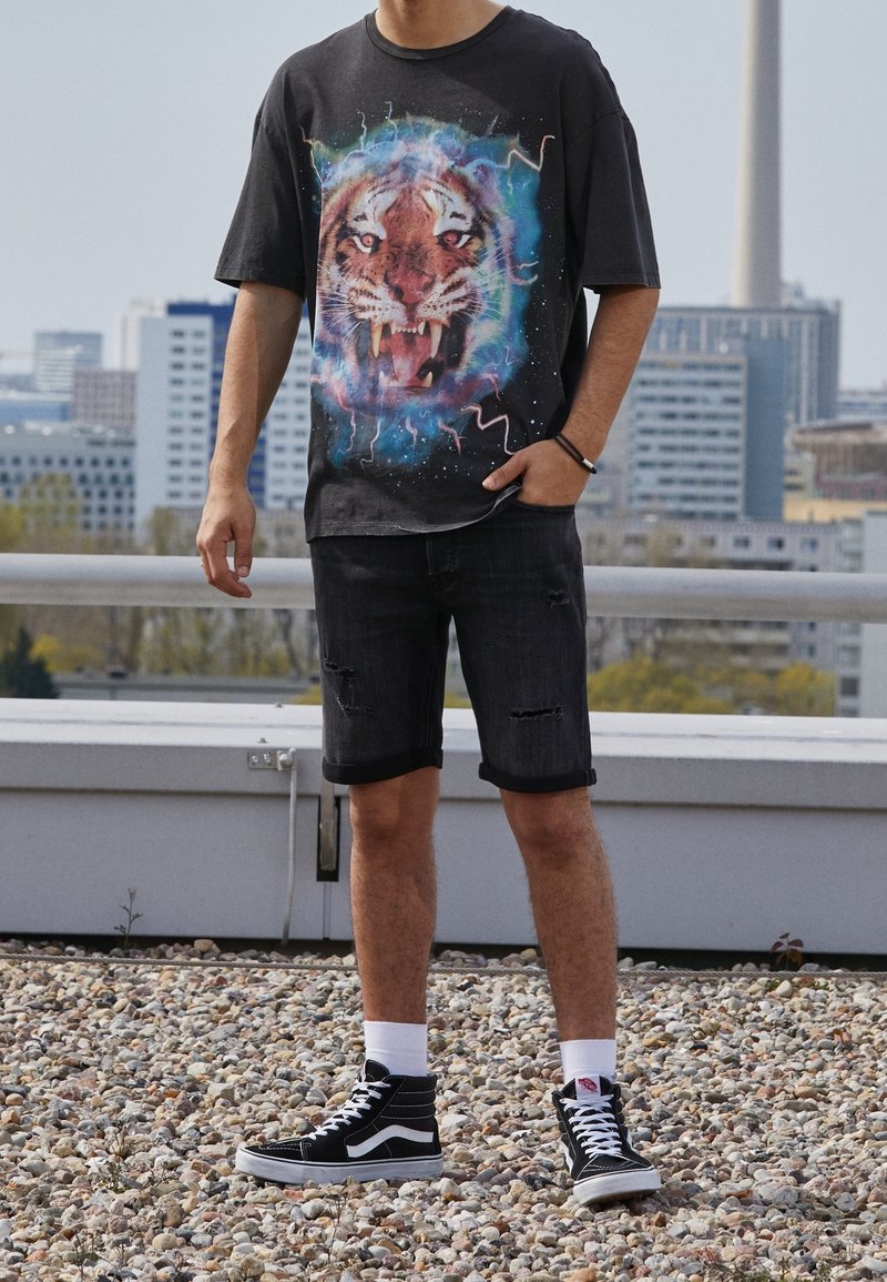 Camiseta gráfica negra con una gran cara de tigre y un fondo de remolinos coloridos; combinada con unos pantalones cortos de mezclilla negra desgastados y unas zapatillas altas negras.