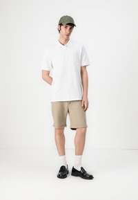 Weißes Poloshirt, beigefarbene Shorts, dunkle Schuhe und grüne Kappe. Das Modell trägt weiße Socken und steht vor einem schlichten Hintergrund. Einfache und legere Kleidung.
