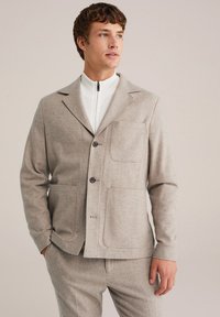 Svetlo bež blazer z teksturo, z ovratnikom tipa notch, tremi gumbi in dvema sprednjima žepoma. Nosi se preko bele jakne z zadrgo. Prilagojen kroj.