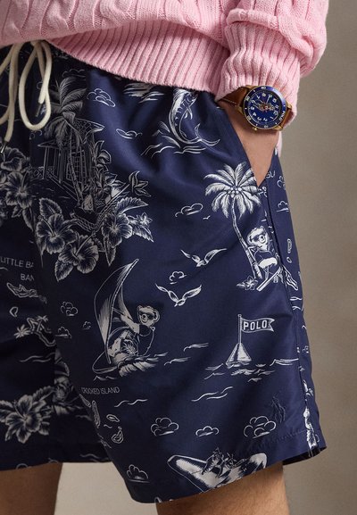 Marineblauwe zwemshorts met witte tropische en nautische illustraties, gedragen met een roze trui, met een polshorloge met een blauwe wijzerplaat en een bruine band.