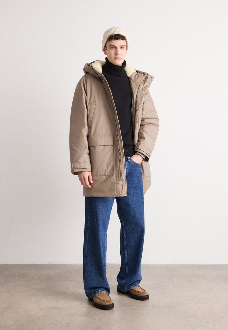 Tan pufferjack met capuchon, crème voering, twee voorzakken, gecombineerd met een zwarte coltrui, brede blauwe jeans en beige instappers.