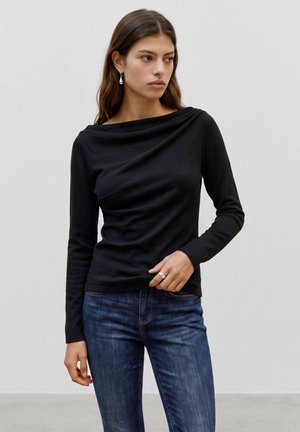 Vrouw met lang bruin haar draagt een zwarte top met lange mouwen, blauwe spijkerbroek, een ring en hangende oorbellen, staand tegen een effen lichte achtergrond.