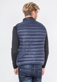 Gilet matelassé bleu marine avec un quilting horizontal, col montant et design sans manches. Porté par-dessus une chemise noire à manches longues.