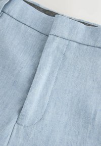 Gros plan sur un tissu en denim bleu clair montrant une ceinture et un détail de braguette zippée cachée sur un pantalon.