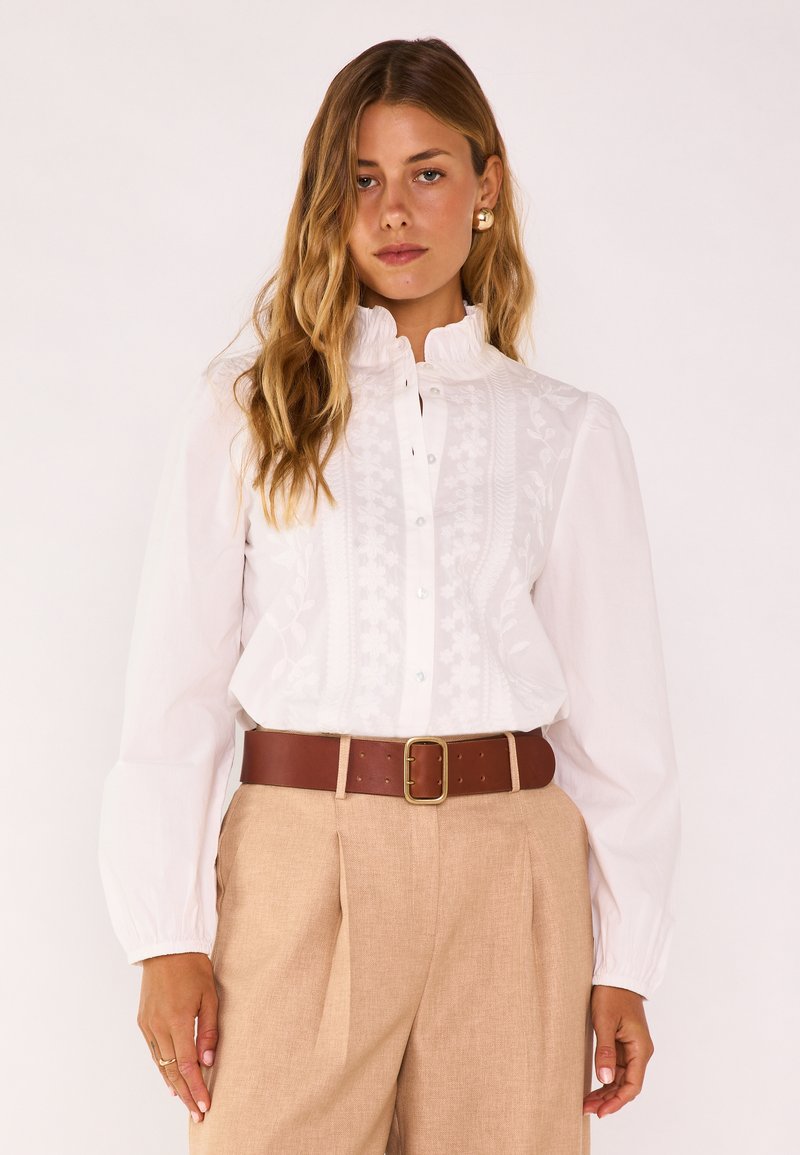 Femme aux cheveux longs et ondulés portant un chemisier blanc brodé à col montant, un pantalon beige, une large ceinture marron et des boucles d'oreilles créoles en or.