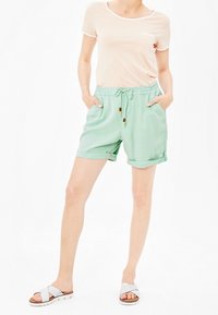 Shorts vert menthe avec une ceinture élastique et des poches latérales, associés à un t-shirt rose clair avec une bordure blanche. Des sandales argentées complètent le look.