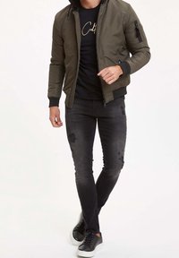 Giubbotto bomber verde oliva con cappuccio, T-shirt nera, jeans neri strappati, e sneakers nere con suole bianche. Dettagli di design minimalisti.