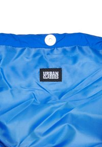 Niebieska tkanina z czarną prostokątną etykieta z napisem "Urban Classics" w białym kolorze, zwieńczona białym napem na niebieskim brzegu.