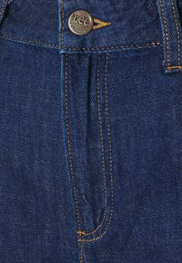 Mörkblå denimjeans med en framknapp med "Lee"-gravyri, kontrasterande gul söm och en lätt mönstrad textur.