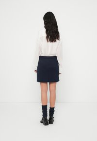 Σκούρα μπλε φούστα με εφαρμοστή μέση, συνδυασμένη με λευκό, φαρδύ πουκάμισο. Το μοντέλο φοράει χοντρές, σκουρόχρωμες κάλτσες και μαύρα παπούτσια με χοντρό τακούνι.