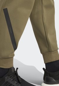 Calças jogger até ao joelho, de cor verde-azeitona, com uma faixa vertical preta e uma textura de cinta contrastante, combinadas com um sapatilho de rede preto.