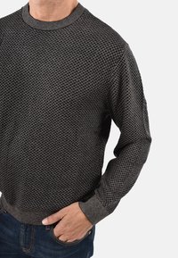 Maglione scuro con motivo a diamante strutturato, caratterizzato da scollo rotondo e polsini a coste. Abbinato a jeans di denim blu.
