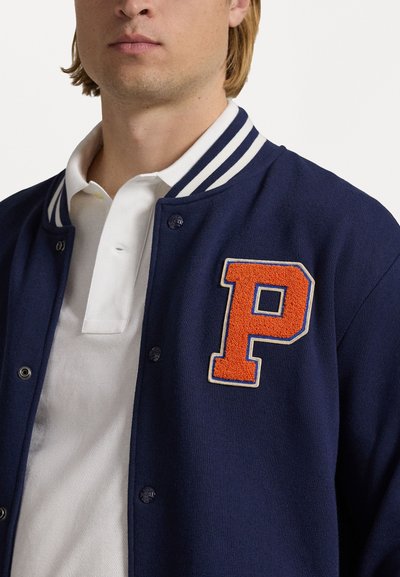 Navy blue varsity jacket with white striped collar and an orange fabric letter "P" patch. Soft texture, button closure, layered over a white polo.