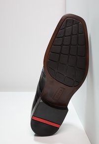 Černá podrážka boty s texturovaným gumovým povrchem, zdobená hnědým okrajem a červeným akcentem na patě. Design je elegantní a moderní.