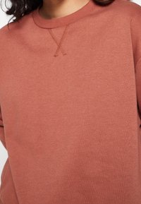 Sudadera de color óxido con cuello redondo, que presenta un detalle texturizado de punto en X en el cuello. Fabricada con una tela suave, de textura lisa.