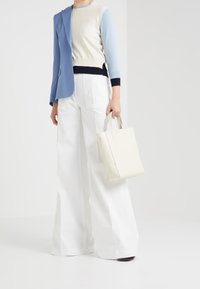 Kvinna i vida vita byxor, en färgblockad tröja, en blå blazer slängd över ena axeln, hållande en vit tote-väska.