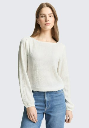 Młoda kobieta ubrana w białą, teksturowaną bluzkę z długim rękawem i niebieskie dżinsy, stojąca na tle jednolitego, jasnoszarego tła.