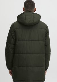 Mørkegrønn polstret kåpe med hette, med diagonal søm, avslappet passform og elastiske ermer. Glatt tekstur, knelengde design.